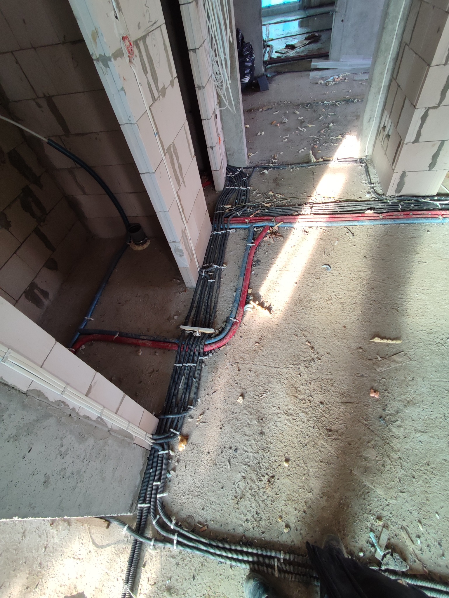 Wiązki przewodów elektrycznych w peszlach, poprowadzone wzdłuż ścian w surowym stanie budynku, z widocznymi puszkami instalacyjnymi i przewodami w kolorach czerwonym i niebieskim.