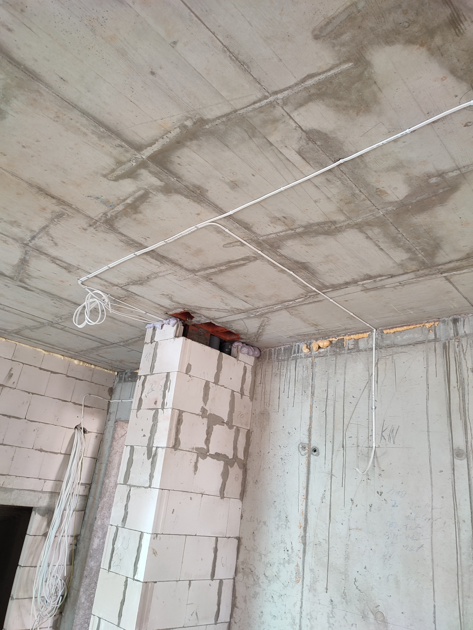 Instalacja elektryczna natynkowa w surowym wnętrzu, widoczne białe kable prowadzone po betonowym suficie i ścianach z pustaków, przyłącze kominowe zabezpieczone pianką montażową.