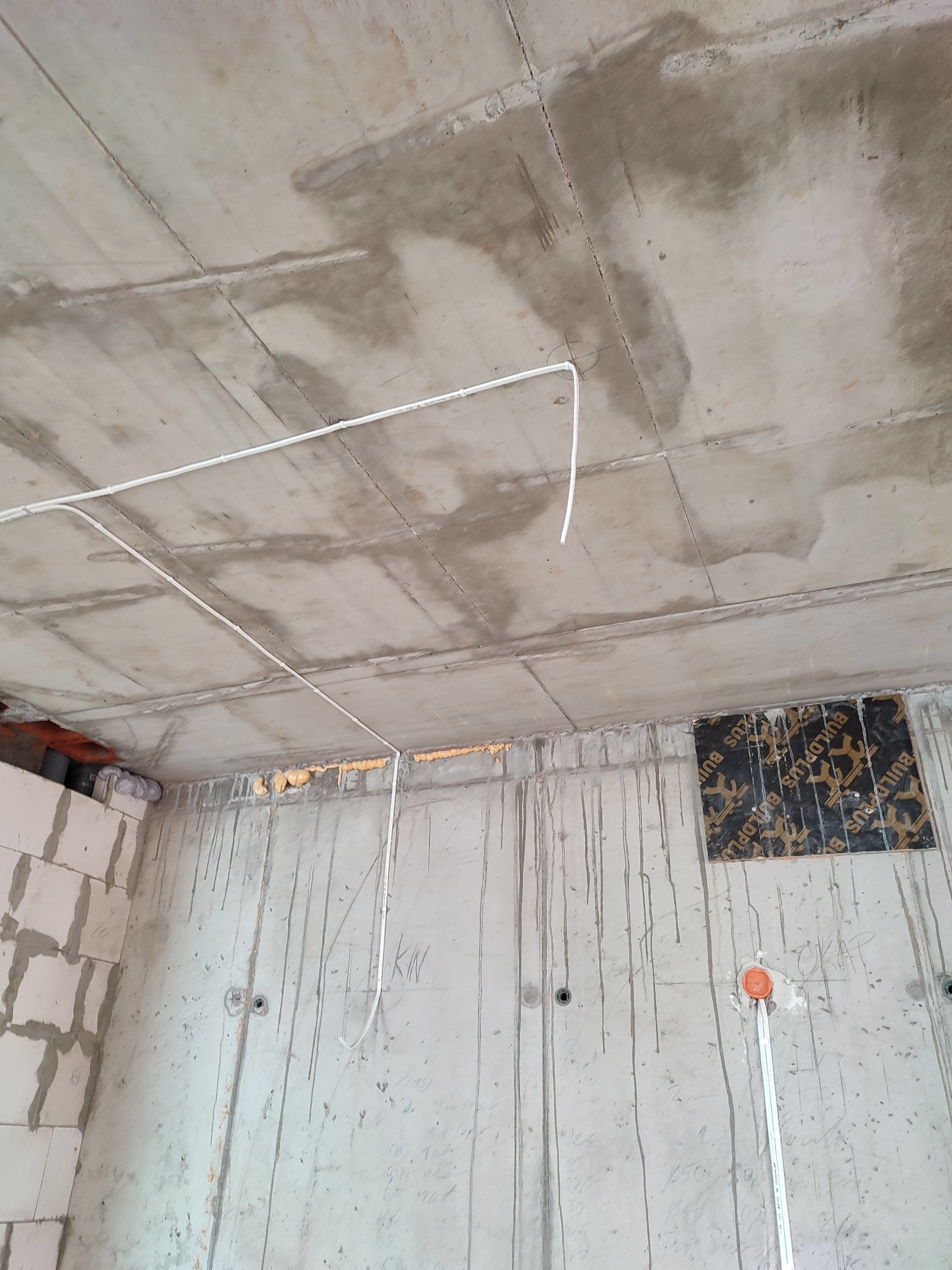 Surowy betonowy strop z widocznymi przewodami elektrycznymi, częściowo zamaskowany otwór w ścianie, ślady po zalaniach i notatki na ścianie.