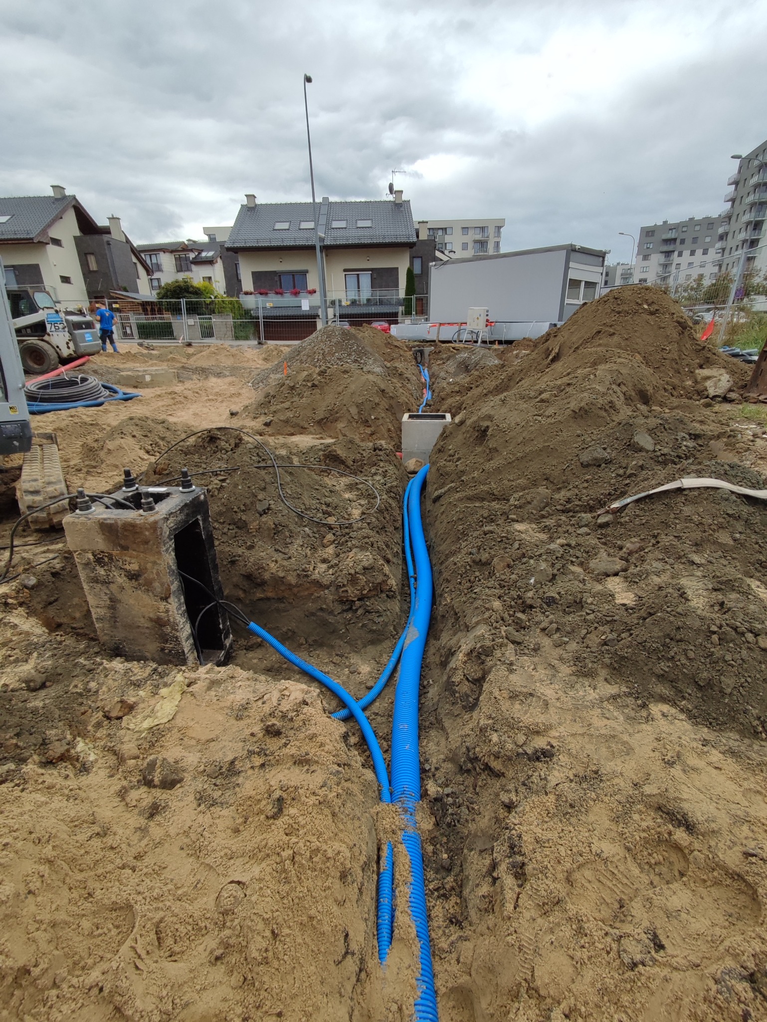 Wykop z widocznymi niebieskimi rurami karbowanymi, prawdopodobnie osłonowymi, prowadzącymi do betonowej studzienki z kablami. W tle widoczne budynki mieszkalne i maszyny budowlane.