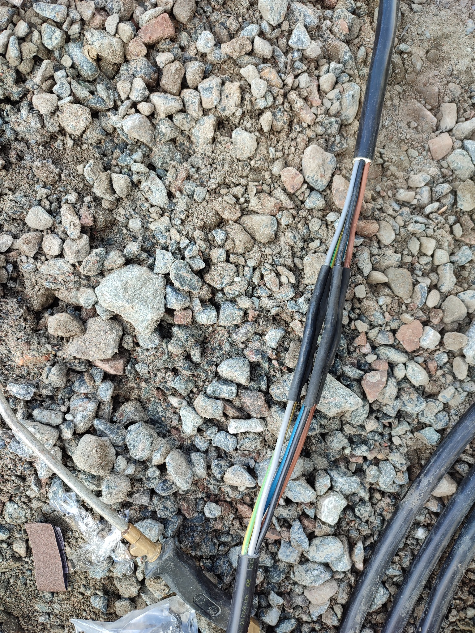 Odsłonięte przewody elektryczne z kolorowymi izolacjami, częściowo zabezpieczone czarną taśmą izolacyjną, leżą na żwirowym podłożu, obok widoczny element instalacji elektrycznej z brązową nakładką.