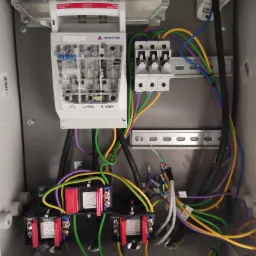 Wnętrze skrzynki elektrycznej z zamontowanym rozłącznikiem bezpiecznikowym RBK00 firmy Apator, transformatorami prądowymi, bezpiecznikami i kolorowymi przewodami.