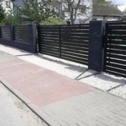 Nowoczesne, poziome, czarne ogrodzenie panelowe z bramą wjazdową, osadzone na betonowych słupkach, z białym żwirem u podstawy, widoczne z poziomu chodnika wyłożonego kostką.