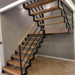 Nowoczesne schody z drewnianymi stopniami i czarną, metalową konstrukcją w minimalistycznym wnętrzu z parkietem ułożonym w jodełkę.