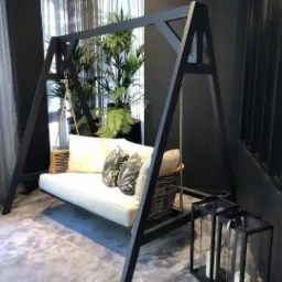 Nowoczesna huśtawka ogrodowa z czarną ramą, wiklinowym siedziskiem, białymi poduszkami i dekoracyjnymi lampionami, ustawiona na tle ciemnego wnętrza z roślinami doniczkowymi.