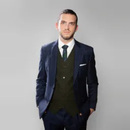 Portret mężczyzny w granatowym garniturze w kratę, zielonej kamizelce i krawacie, z rękami w kieszeniach, na jasnoszarym tle.