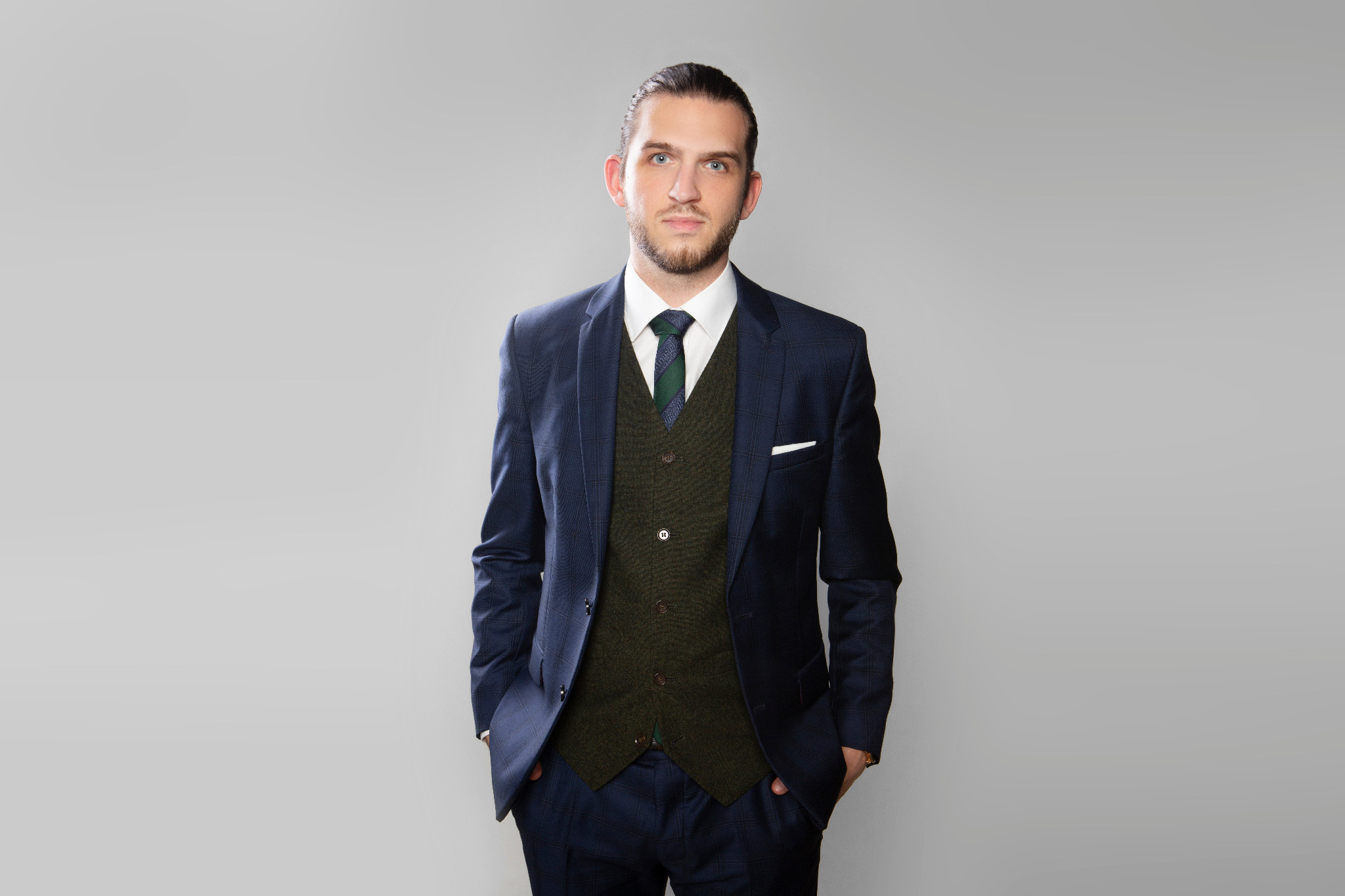 Portret mężczyzny w granatowym garniturze w kratę, zielonej kamizelce i krawacie, z rękami w kieszeniach, na jasnoszarym tle.