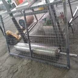 Metalowa klatka z siatki z widocznym w środku cylindrem, prawdopodobnie element maszyny, w warsztacie.