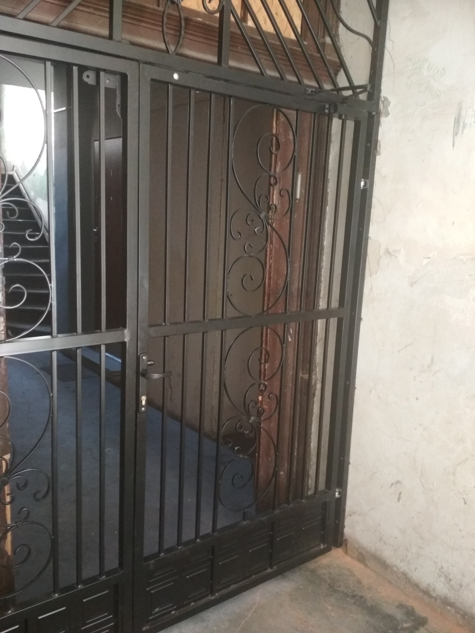 Czarna, metalowa krata drzwiowa z ozdobnymi, zakręconymi elementami, zabezpieczająca wejście do budynku z widocznymi schodami w tle.