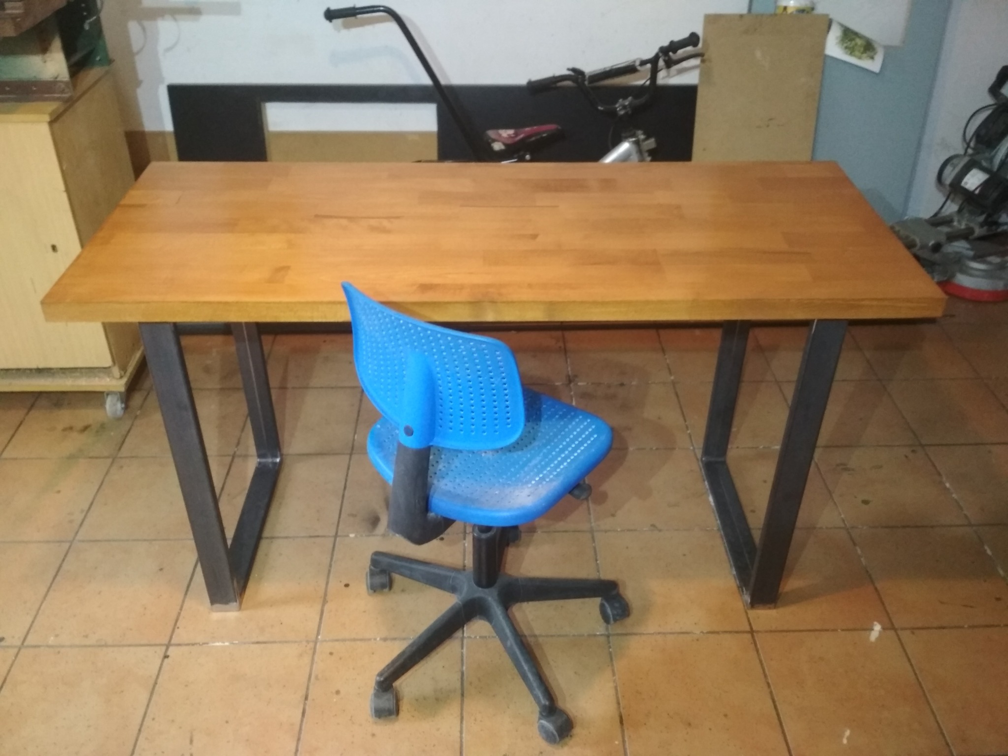 Biurko z blatem z jasnego drewna i nogami z profili stalowych, na podłodze z terakoty, z niebieskim krzesłem obrotowym, w tle widoczne narzędzia i materiały.