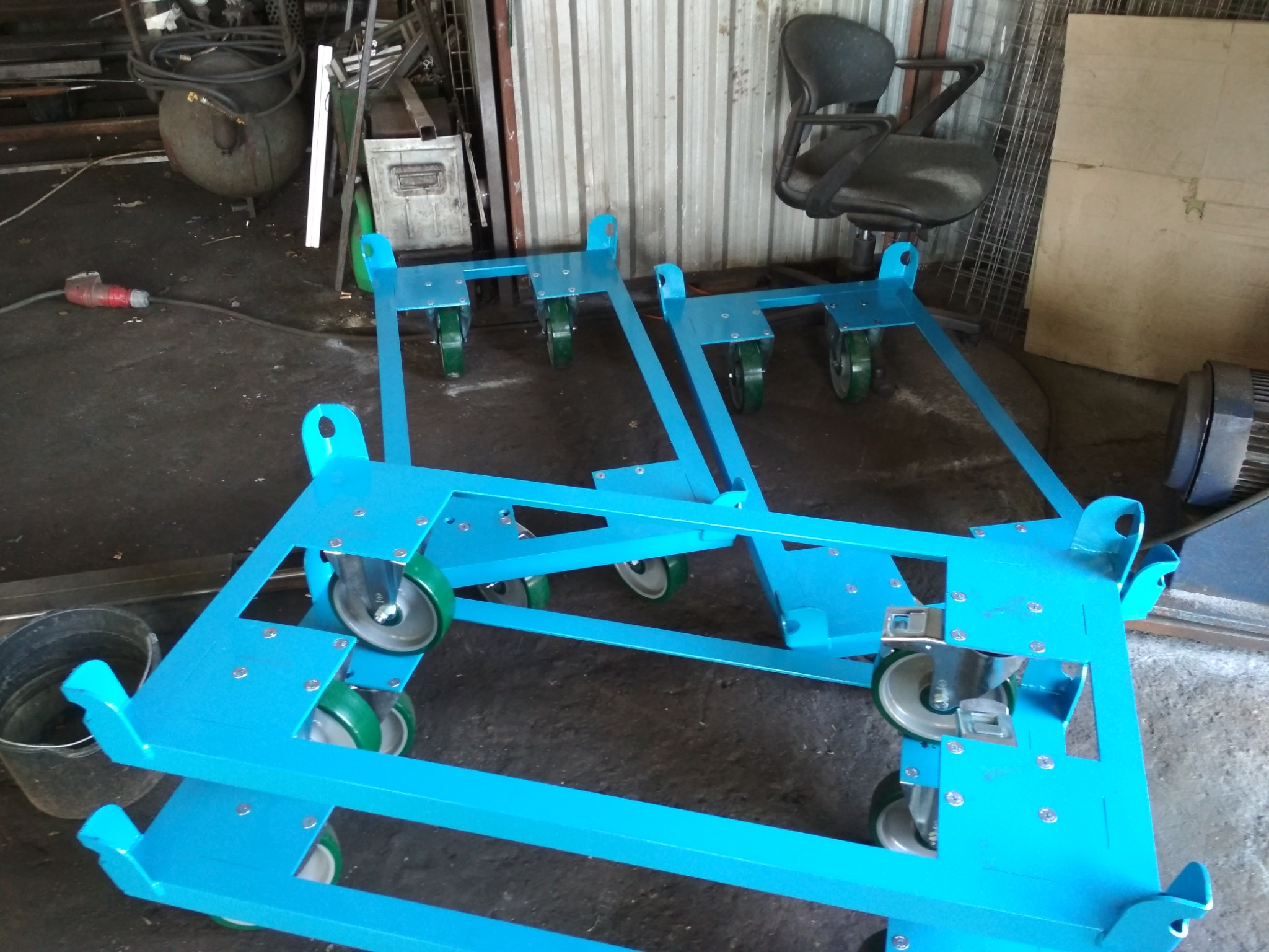 Dwa wózki transportowe z niebieskiej stali z zielonymi kółkami, stojące w warsztacie, widoczne spawane łączenia i śruby mocujące.