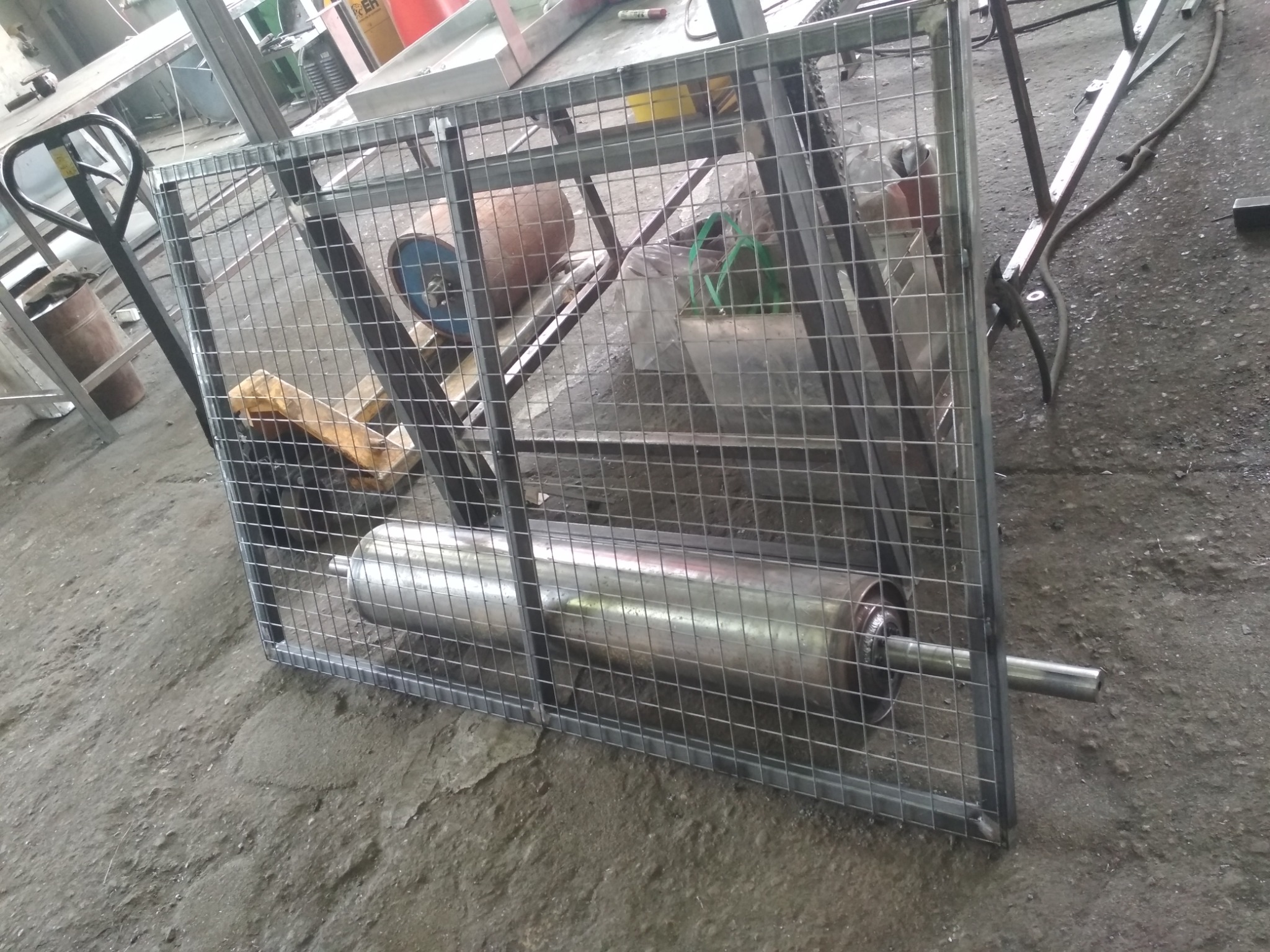 Metalowa klatka z siatki z widocznym w środku cylindrem, prawdopodobnie element maszyny, w warsztacie.