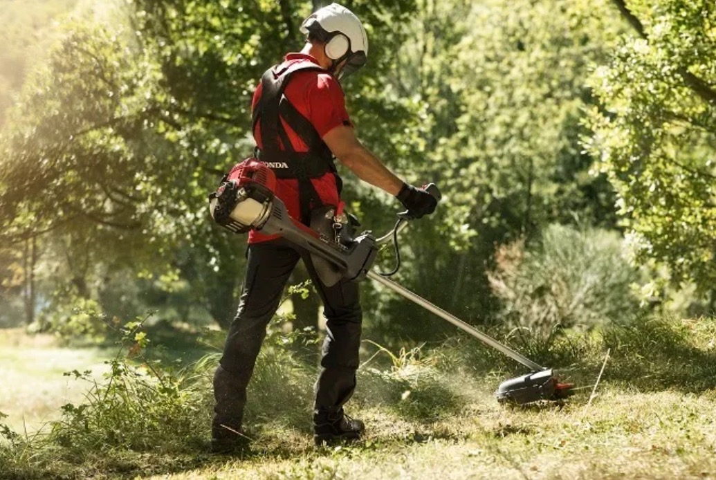 Pracownik w kasku i uprzęży, z podkaszarką Honda, przycina trawę na nierównym terenie w słoneczny dzień. W tle widoczne drzewa i krzewy.