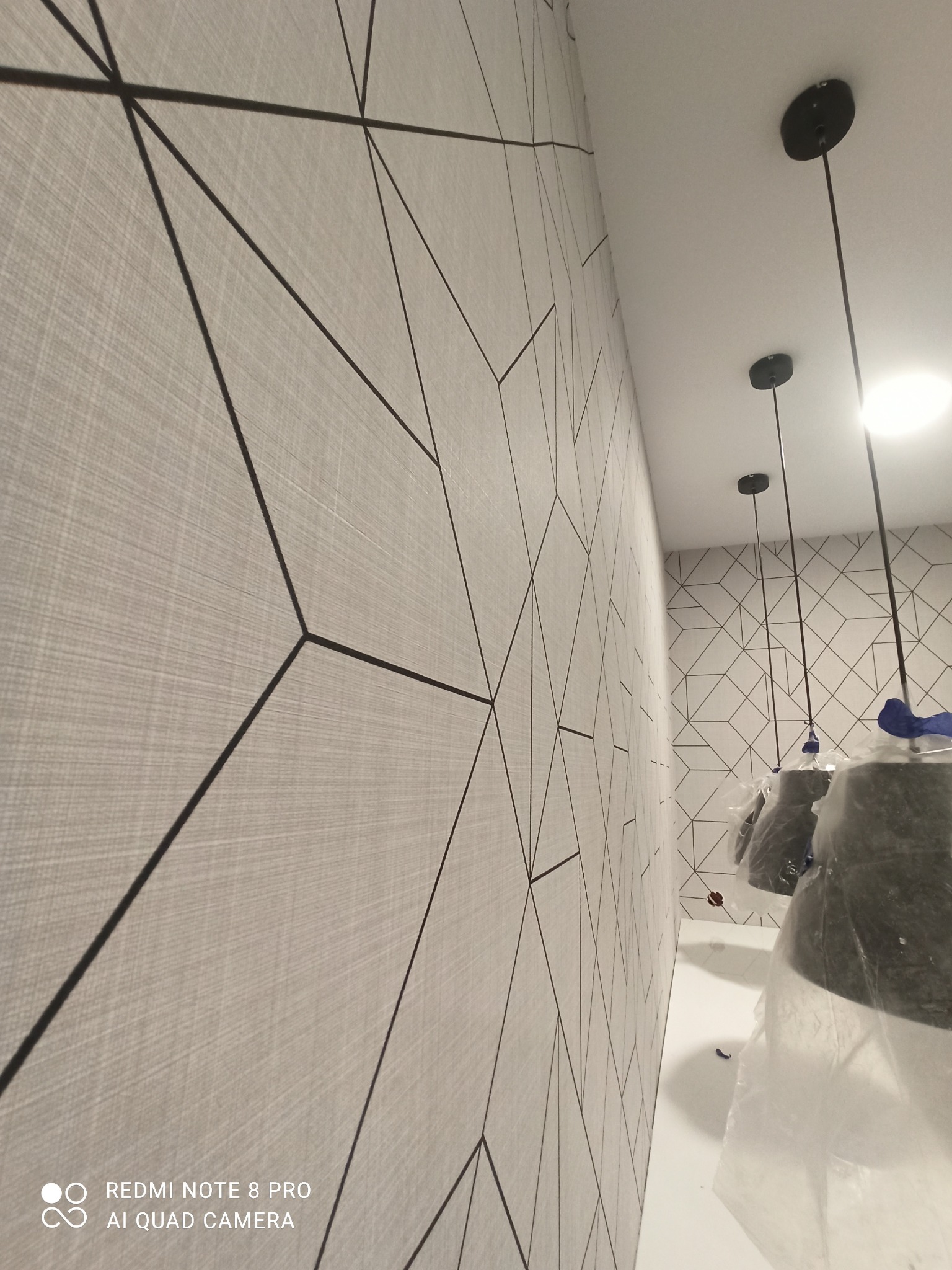 Ściana pokryta jasną tapetą z geometrycznym wzorem czarnych linii, widoczne lampy wiszące zabezpieczone folią ochronną.
