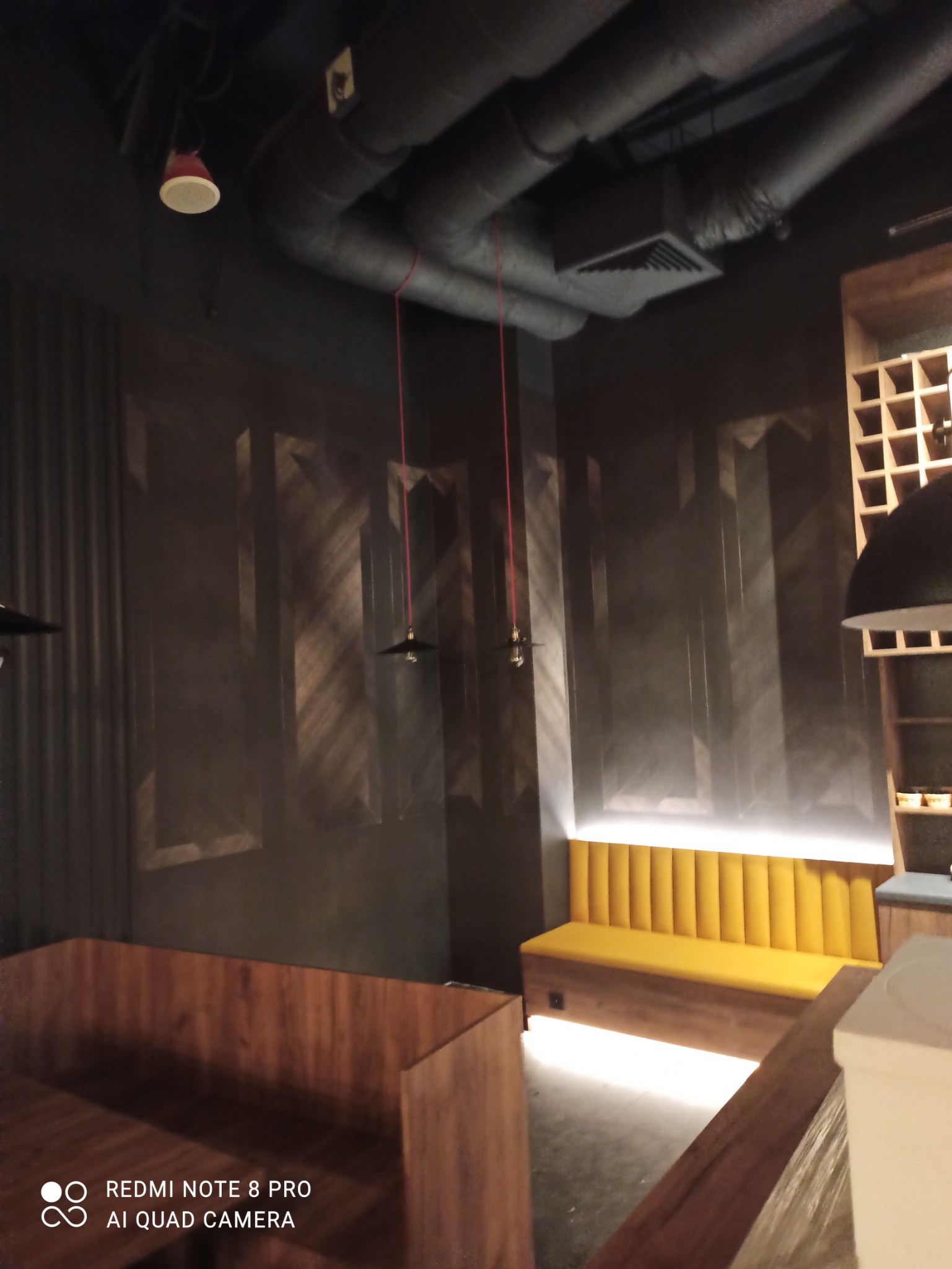 Ciemne wnętrze lokalu gastronomicznego z tapetą w geometryczne wzory, żółtą tapicerowaną ławką podświetloną od dołu oraz drewnianymi boksami.