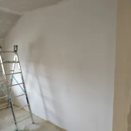 Ściana po nałożeniu gładzi, widoczna drabina aluminiowa, sufit w trakcie prac wykończeniowych, podłoga zabezpieczona folią.