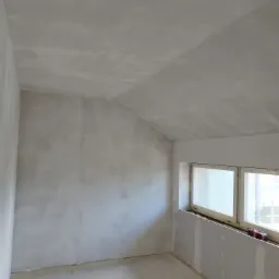 Surowe wnętrze w trakcie prac wykończeniowych: ściany i sufit po nałożeniu szpachli, widoczne ślady po łatach, okno z parapetem, na którym leży czerwona skrzynka narzędziowa.