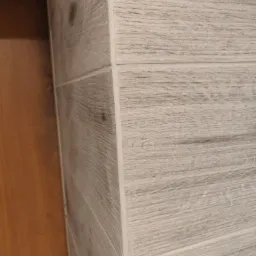 Fragment wykończenia wnętrza: narożnik obłożony płytkami imitującymi drewno w odcieniach szarości, przylegający do brązowych drzwi.