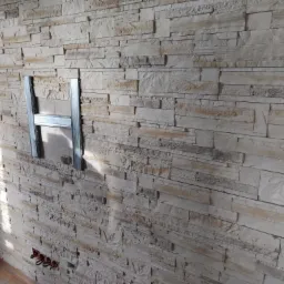 Ściana wykończona dekoracyjną okładziną kamienną z zamontowanym metalowym profilem w kształcie litery H, prawdopodobnie przygotowanie do montażu płyty kartonowo-gipsowej.