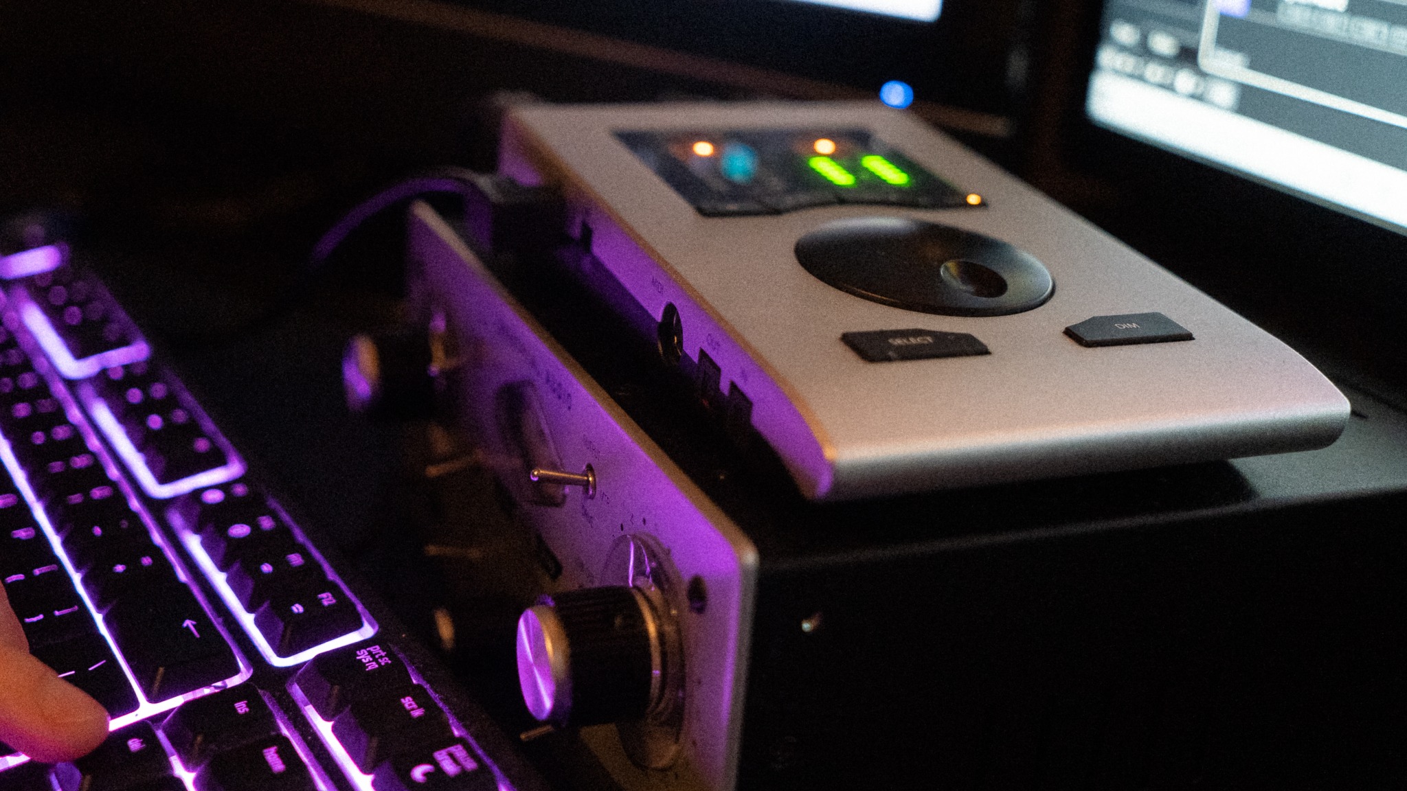 Fragment studia nagraniowego z klawiaturą podświetloną na fioletowo, interfejsem audio z podświetlonymi diodami i wzmacniaczem słuchawkowym z pokrętłem, ukazujący proces tworzenia muzyki...
