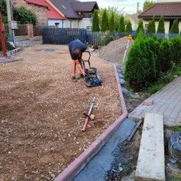Pracownik zagęszcza podłoże pod kostkę brukową za pomocą zagęszczarki spalinowej; widoczne obrzeża, świeżo wylany beton, poziomica, stos iglaków w tle.