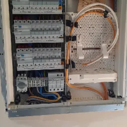 Otwarta skrzynka elektryczna w trakcie montażu, widoczne liczne bezpieczniki, przewody w różnych kolorach, listwa zaciskowa i tabliczka z odręcznymi notatkami na temat podłączeń.