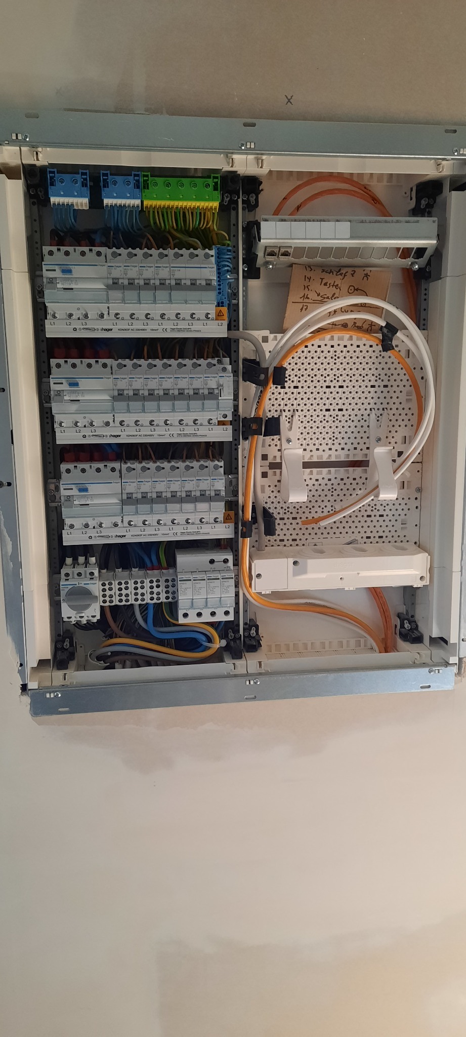 Otwarta skrzynka elektryczna w trakcie montażu, widoczne liczne bezpieczniki, przewody w różnych kolorach, listwa zaciskowa i tabliczka z odręcznymi notatkami na temat podłączeń.