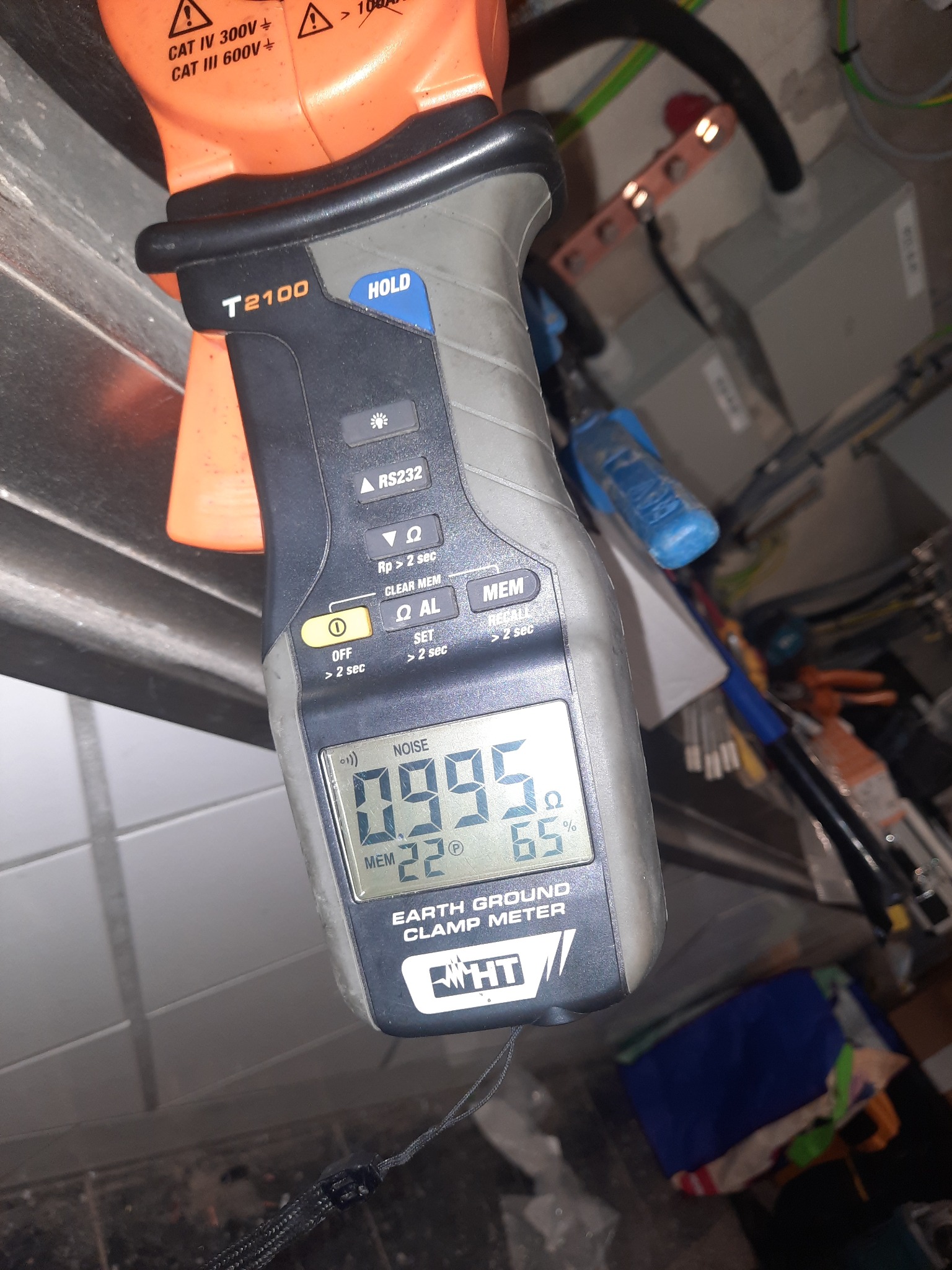 Miernik cęgowy HT T2100 wskazujący wartość rezystancji uziemienia 0.995 oma, używany do weryfikacji instalacji elektrycznej w tle widoczne kable i elementy instalacji.