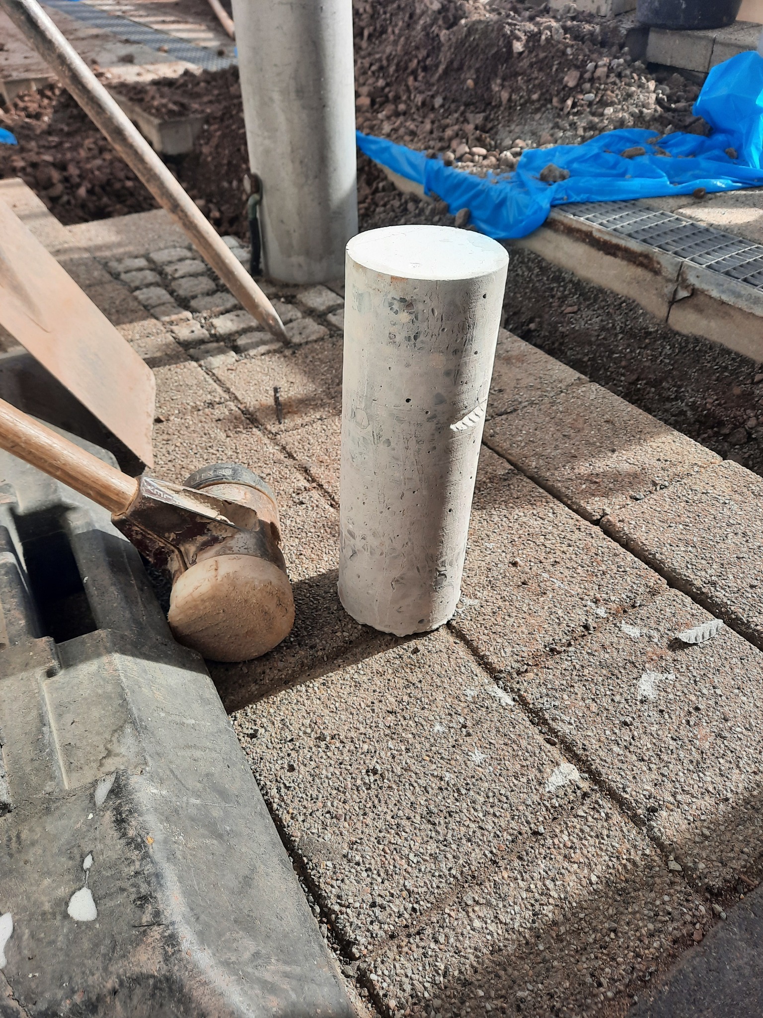 Cylindryczny rdzeń betonowy na kostce brukowej, obok gumowy młotek i łopata, prawdopodobnie pobrany do badań wytrzymałościowych na budowie.