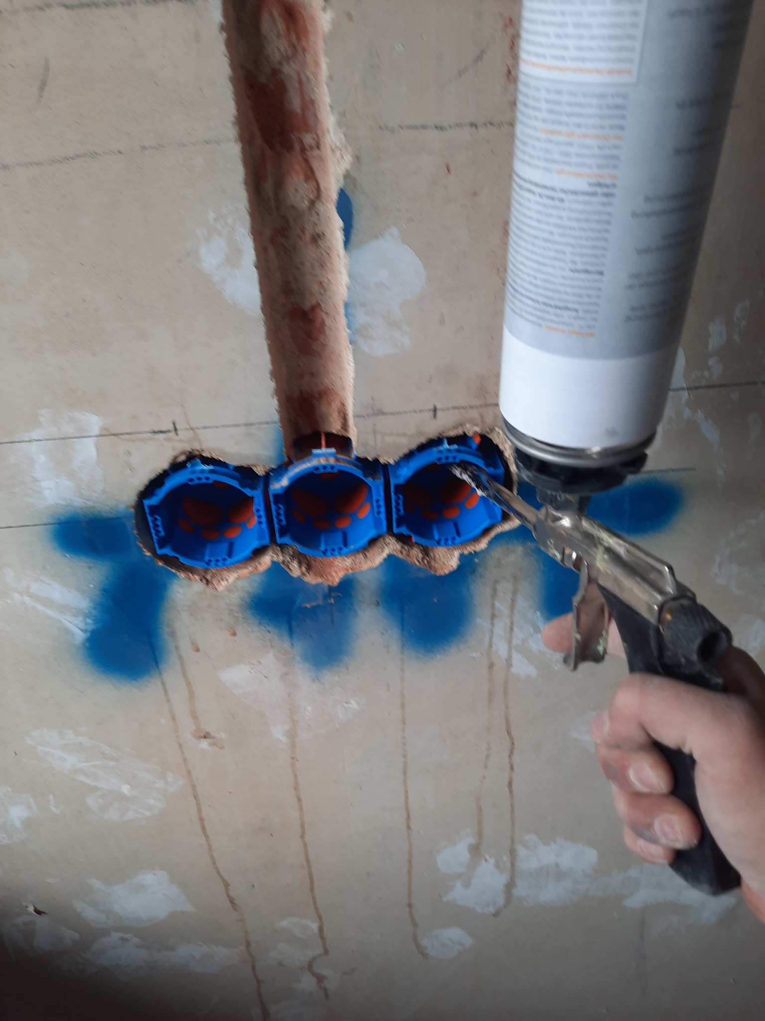 Wypełnianie pianką montażową przestrzeni wokół trzech niebieskich, podwójnych puszek elektrycznych w ścianie, widoczna rura instalacyjna i ślady niebieskiej farby w sprayu.