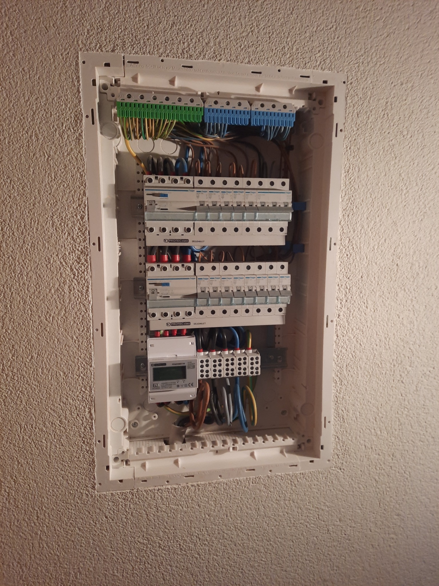 Rozdzielnica elektryczna w białej obudowie, zamontowana na ścianie o fakturze baranka, z widocznymi bezpiecznikami, listwami zaciskowymi i przewodami w różnych kolorach.