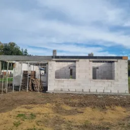 Surowy stan budynku z betonu komórkowego w Nowym Tomyślu, widoczne palety z materiałem budowlanym i metalowe rusztowanie na tle drzew.