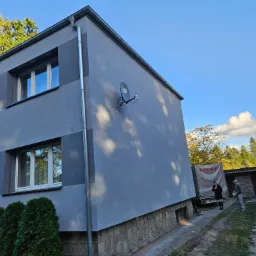 Dwupiętrowy budynek z szarą elewacją i fragmentem kamiennej podmurówki, widoczny z boku, z anteną satelitarną na ścianie i dwiema osobami oraz samochodem dostawczym w tle.