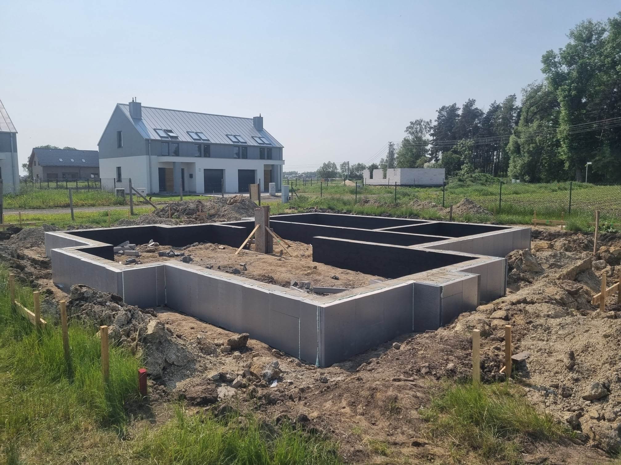 Wylane fundamenty domu jednorodzinnego z widocznym ociepleniem styropianem, w tle dom w trakcie budowy i zieleń.