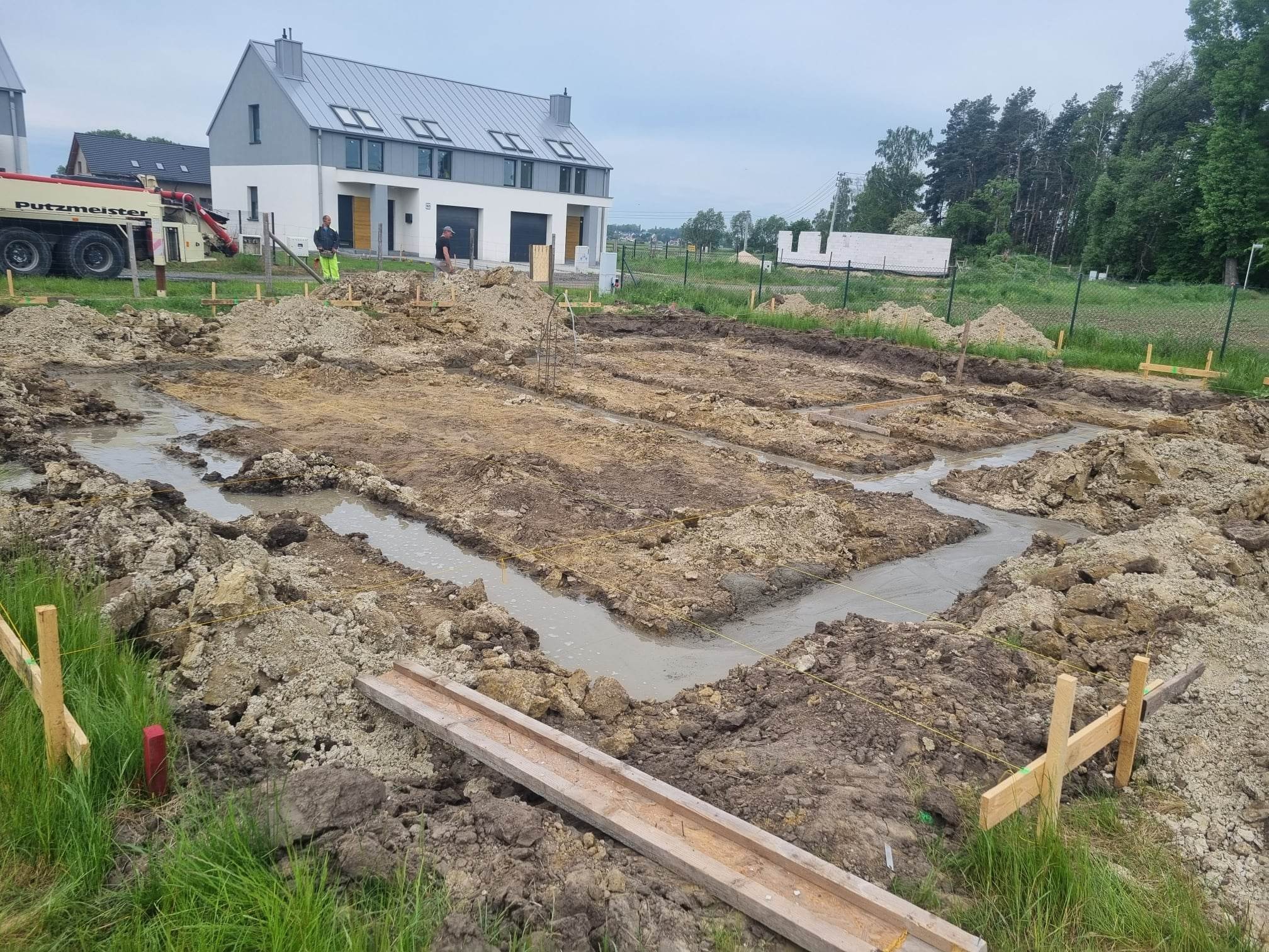 Przygotowanie fundamentów pod budowę domu jednorodzinnego: zalane betonem wykopy, widoczny szalunek, z tyłu nowoczesny dom w trakcie budowy oraz betoniarka Putzmeister.