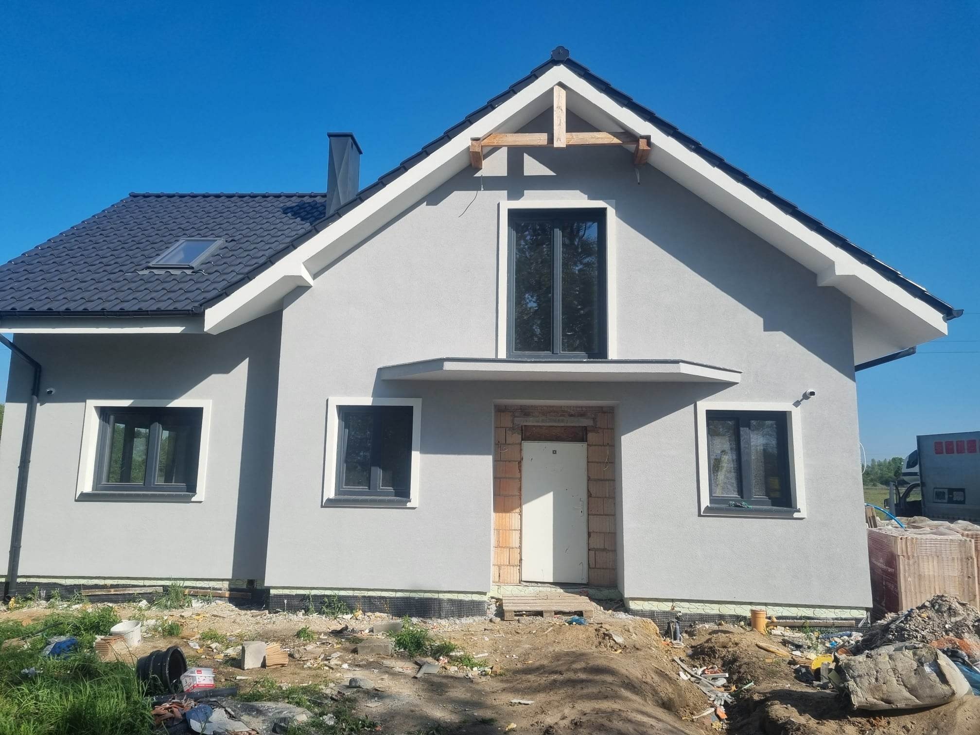 Szary dom jednorodzinny z ciemną dachówką i czarnymi oknami w trakcie budowy, widoczne rusztowania na szczycie dachu i materiały budowlane na ziemi.