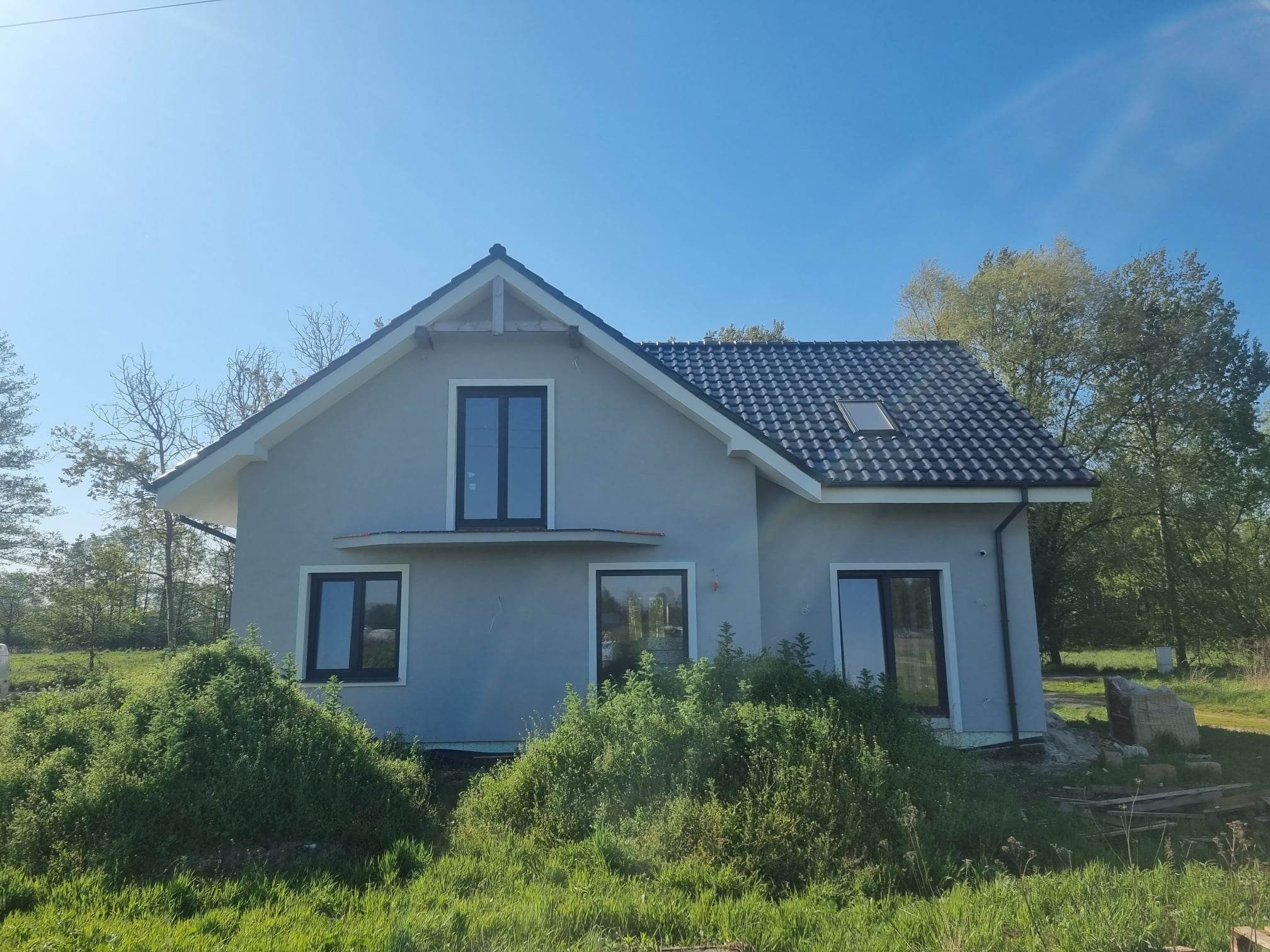 Dom jednorodzinny z szarą elewacją, czarną dachówką i oknami, częściowo zasłonięty bujną zielenią, sfotografowany w słoneczny dzień.