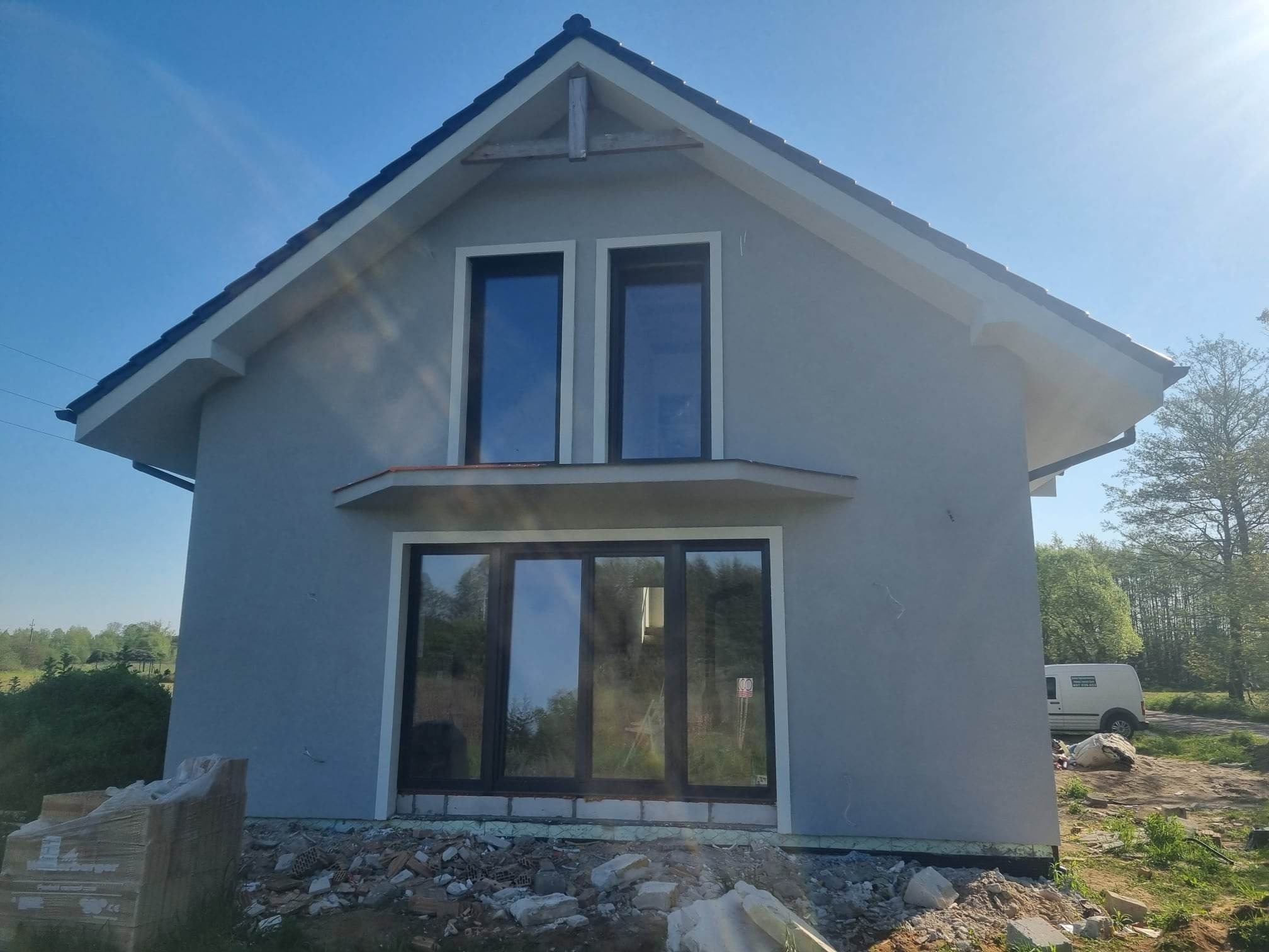 Dom jednorodzinny z szarą elewacją po nałożeniu tynku strukturalnego, widoczne okna z ciemnymi ramami, teren wokół domu w trakcie prac wykończeniowych, sterta materiałów budowlanych obok.