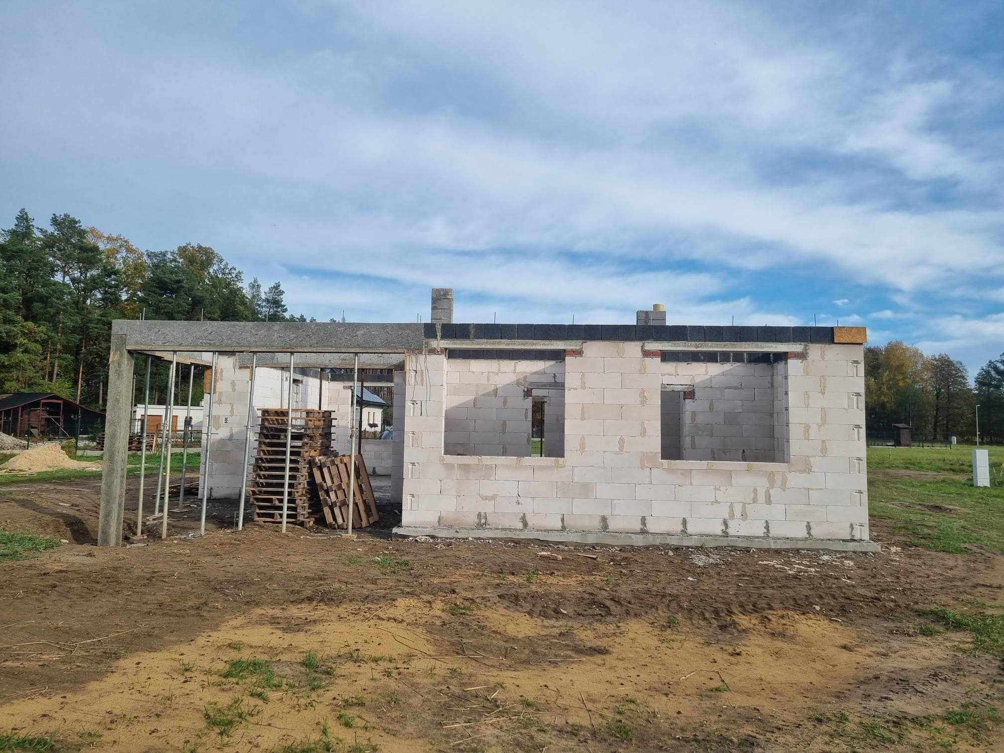 Surowy stan budynku z betonu komórkowego w Nowym Tomyślu, widoczne palety z materiałem budowlanym i metalowe rusztowanie na tle drzew.