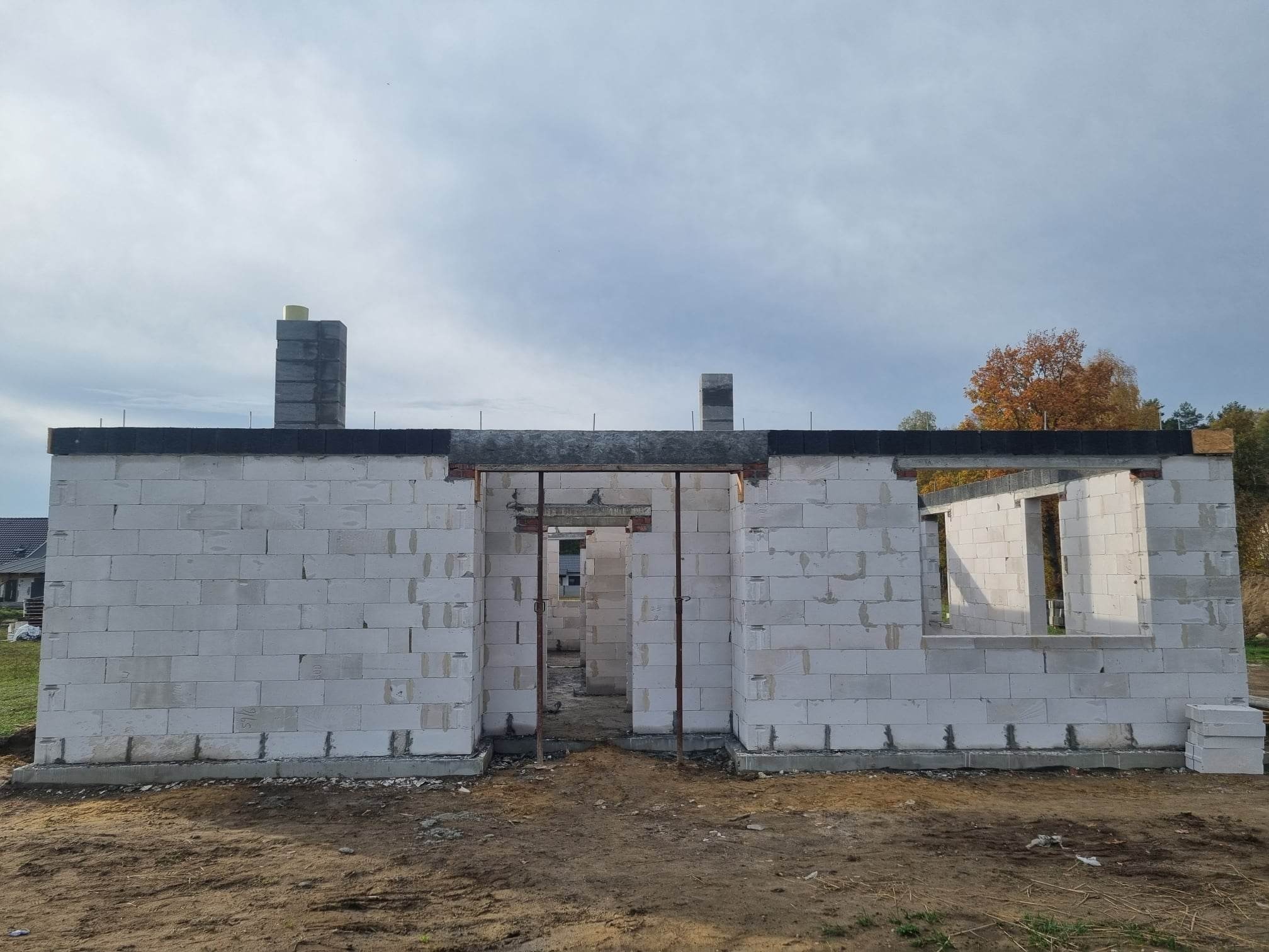 Surowy stan ścian zewnętrznych budynku z bloczków betonowych, widoczne otwory okienne i drzwiowe, komin z szarej cegły, teren budowy z ziemią, w tle drzewa i fragment dachu sąsiedniego domu.