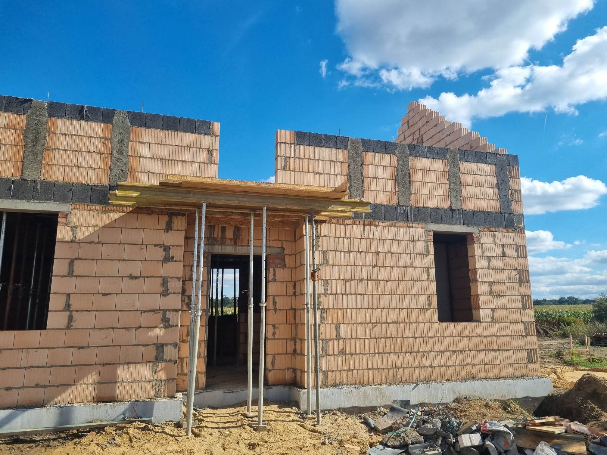 Wznoszenie ścian parterowego domu z bloczków ceramicznych, widoczne otwory okienne i drzwiowe, podpory stropowe, ocieplenie wieńca z czarnego styropianu.
