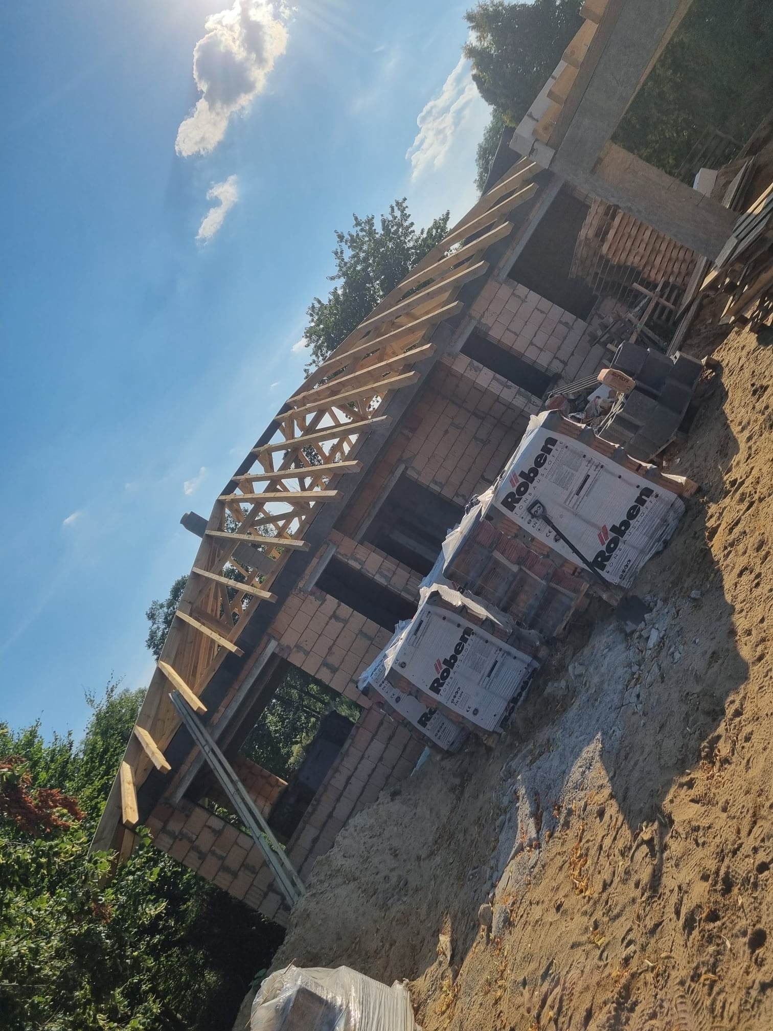 Wznoszenie ścian z cegły klinkierowej i konstrukcji dachu z drewnianych belek, widoczne pakiety cegieł Roben na placu budowy w słoneczny dzień.