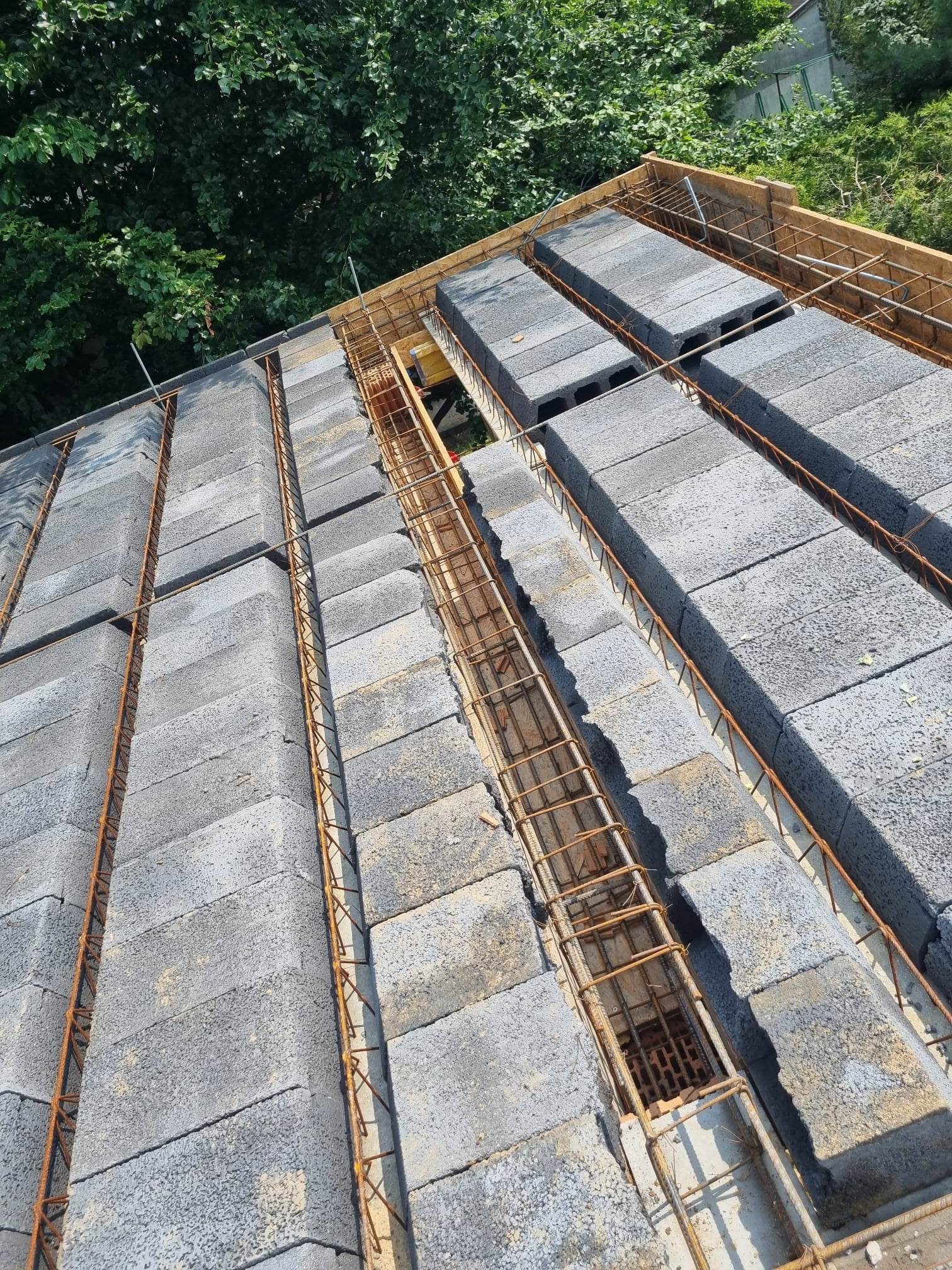 Konstrukcja stropu Teriva z pustaków szalunkowych i zbrojenia stalowego, widok z góry na budowie, z zielenią drzew w tle.