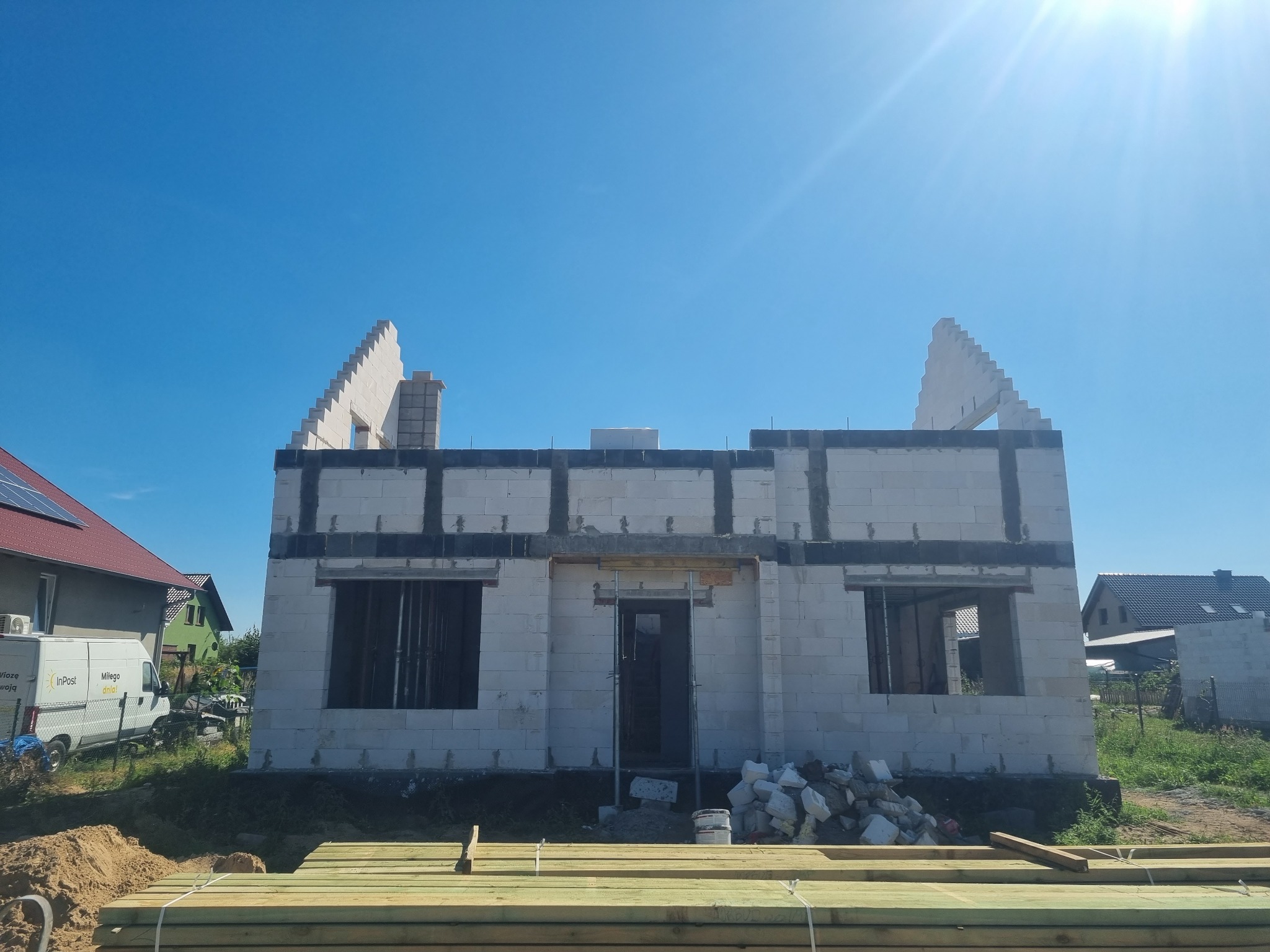 Surowy stan budynku jednorodzinnego z betonu komórkowego, widoczne niezadaszone fragmenty ścian szczytowych, przygotowane otwory okienne i drzwiowe, składowane drewno budowlane na pierwszym planie,...