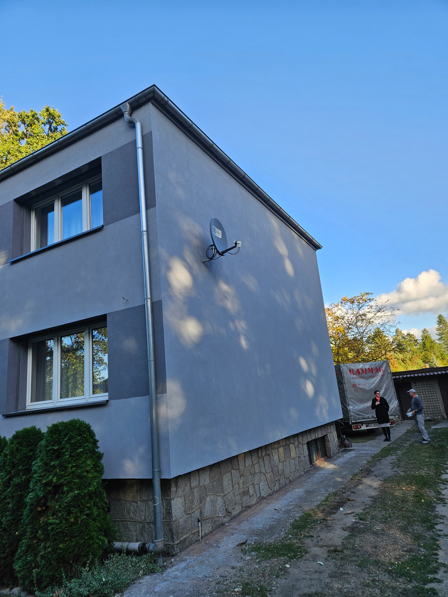 Dwupiętrowy budynek z szarą elewacją i fragmentem kamiennej podmurówki, widoczny z boku, z anteną satelitarną na ścianie i dwiema osobami oraz samochodem dostawczym w tle.