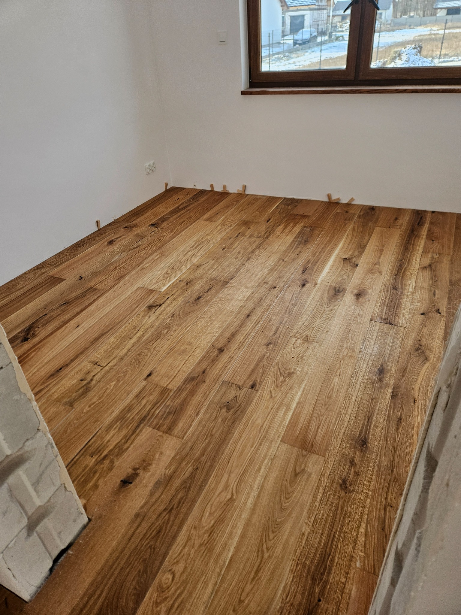 Ułożona podłoga z paneli drewnianych w jasnym odcieniu, z widocznymi klinami dystansowymi przy ścianach i fragmentem okna w tle.