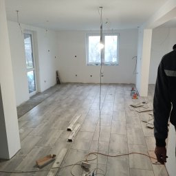 PAWEŁ MOŚCICKI - Układanie podłogi z płytek imitujących drewno w jasnym pomieszczeniu. Widoczne listwy przypodłogowe, kable elektryczne, krzyżyki dystansowe i fragment osoby w ubraniu roboczym.