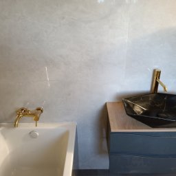 PAWEŁ MOŚCICKI - Wykończona łazienka w Łukowie: czarna, geometryczna umywalka nablatowa z złotą armaturą, biała wanna z złotą baterią na tle szarych, marmurkowych płytek.