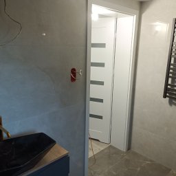 PAWEŁ MOŚCICKI - Nowoczesna łazienka w szarościach z elementami złota, czarna umywalka nablatowa, grzejnik drabinkowy i drzwi z przeszkleniami, widoczne niedokończone prace elektryczne.