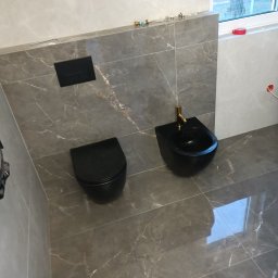 PAWEŁ MOŚCICKI - Wykończona łazienka z szarymi płytkami imitującymi marmur, wisząca czarna toaleta i bidet z złotą armaturą, widoczny grzejnik drabinkowy i okno.